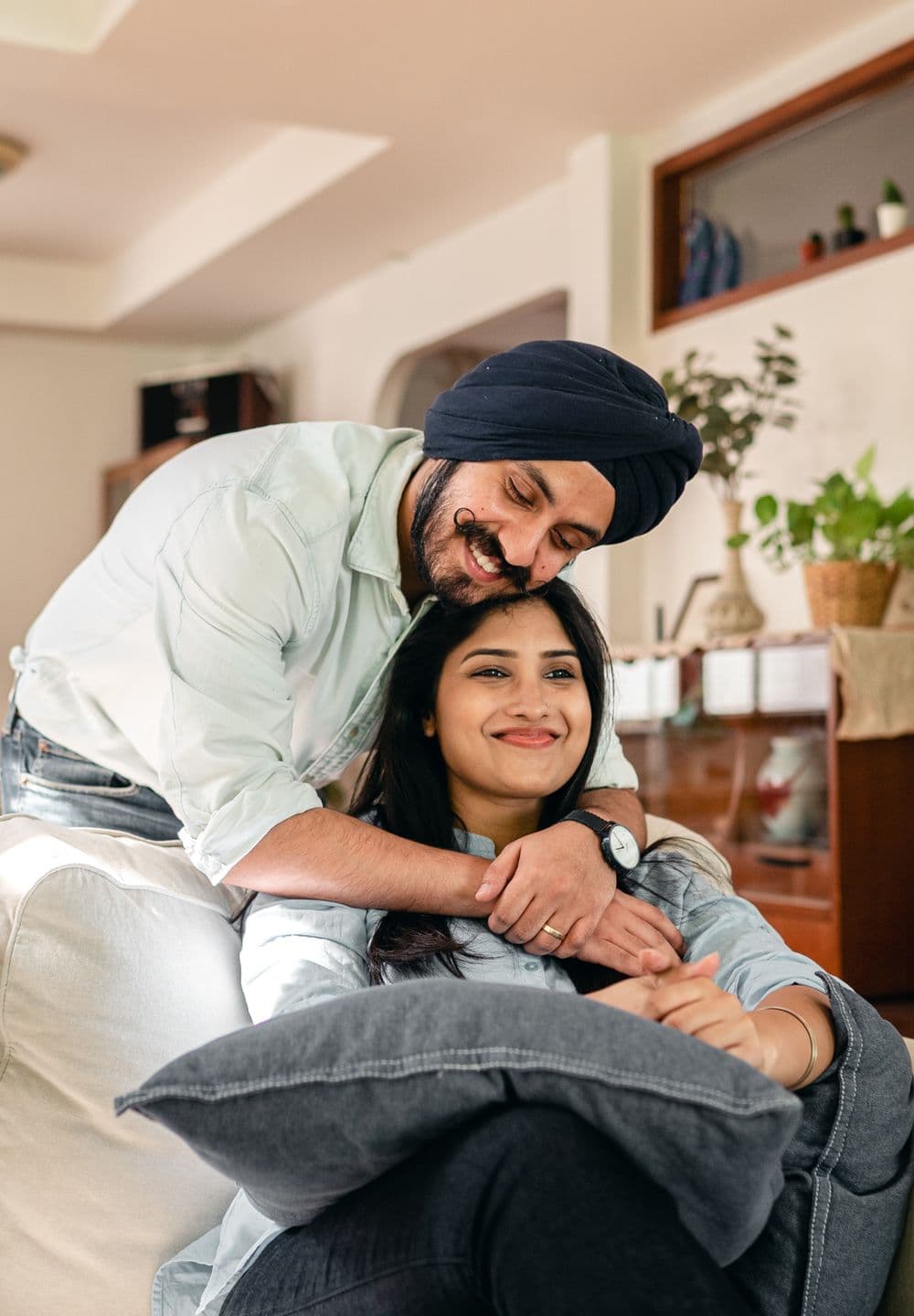 A man hugs his partner from behind on the sofa; both are smiling.
